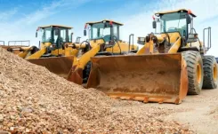 Three yellow excavators lined up against a pile of stones and rubble with blue sky above.